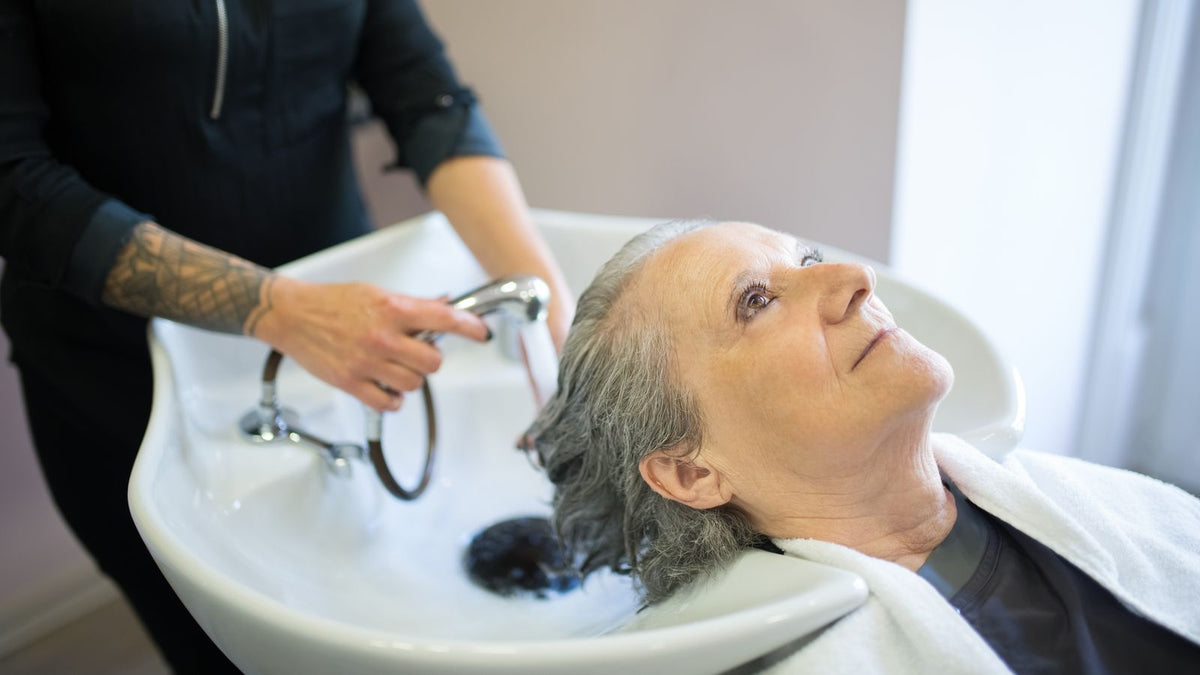 Senior woman getting hair washed by stylist in a cozy salon setting, illustrating hormone therapy hair care