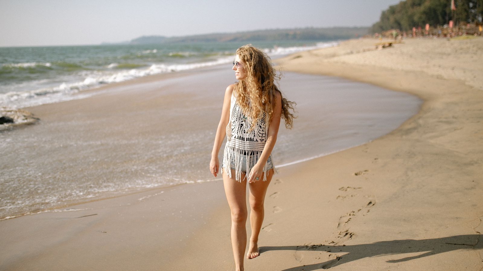 Woman with curly hair walking barefoot on a sunny beach, asking is salt water good for your hair