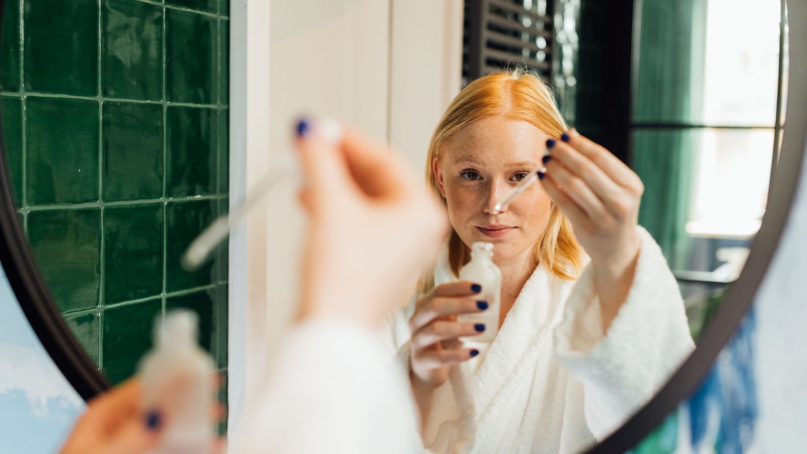 Woman applying serum at a bathroom mirror, following a simple skincare routine order that works