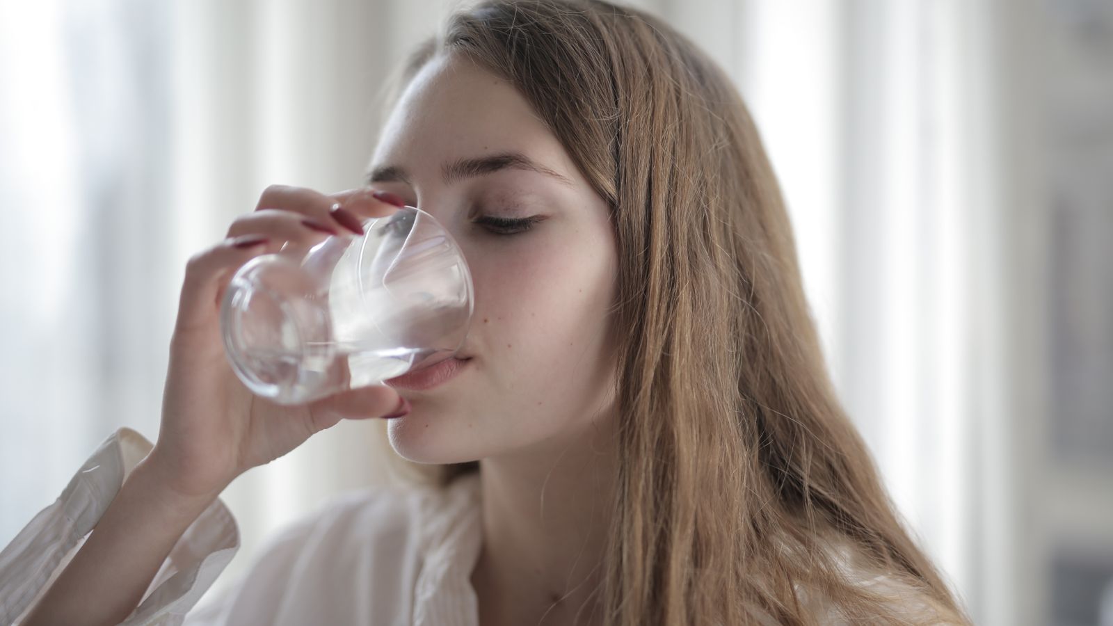 Woman drinking a glass of water indoors, asking does drinking water help hair
