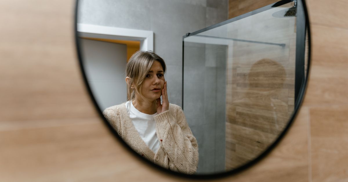 Woman studying her reflection in a bathroom mirror, checking the parting changes linked to hormone replacement hair loss