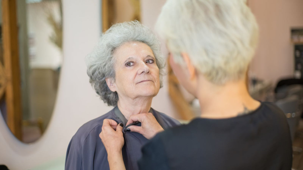 Senior woman getting ready in a hair salon, assisted by a hairstylist adjusting her cape, illustrating hormone therapy hair restoration