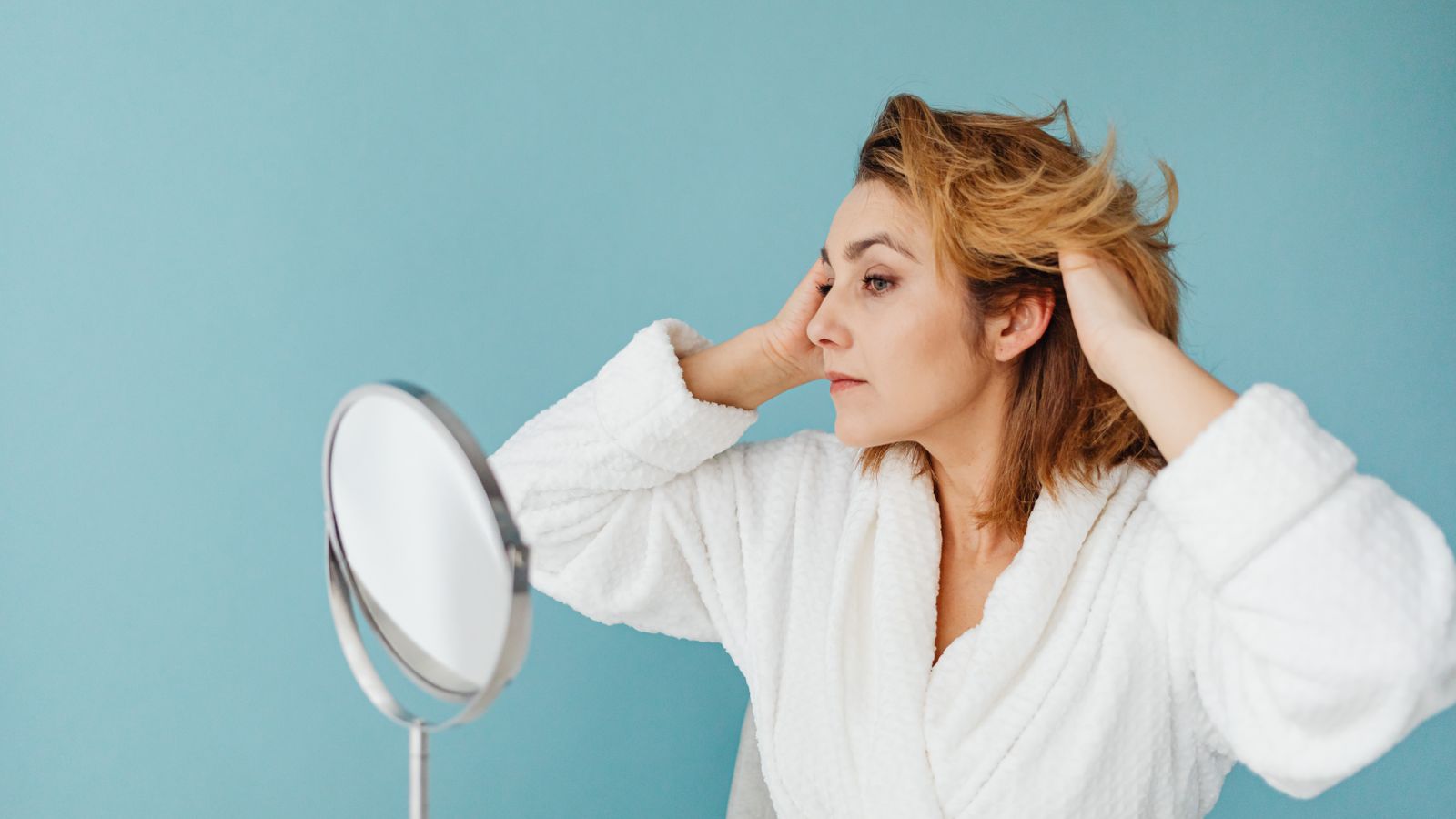 Woman in a bathrobe fixing her hair at a mirror, checking for hormone therapy hairline changes