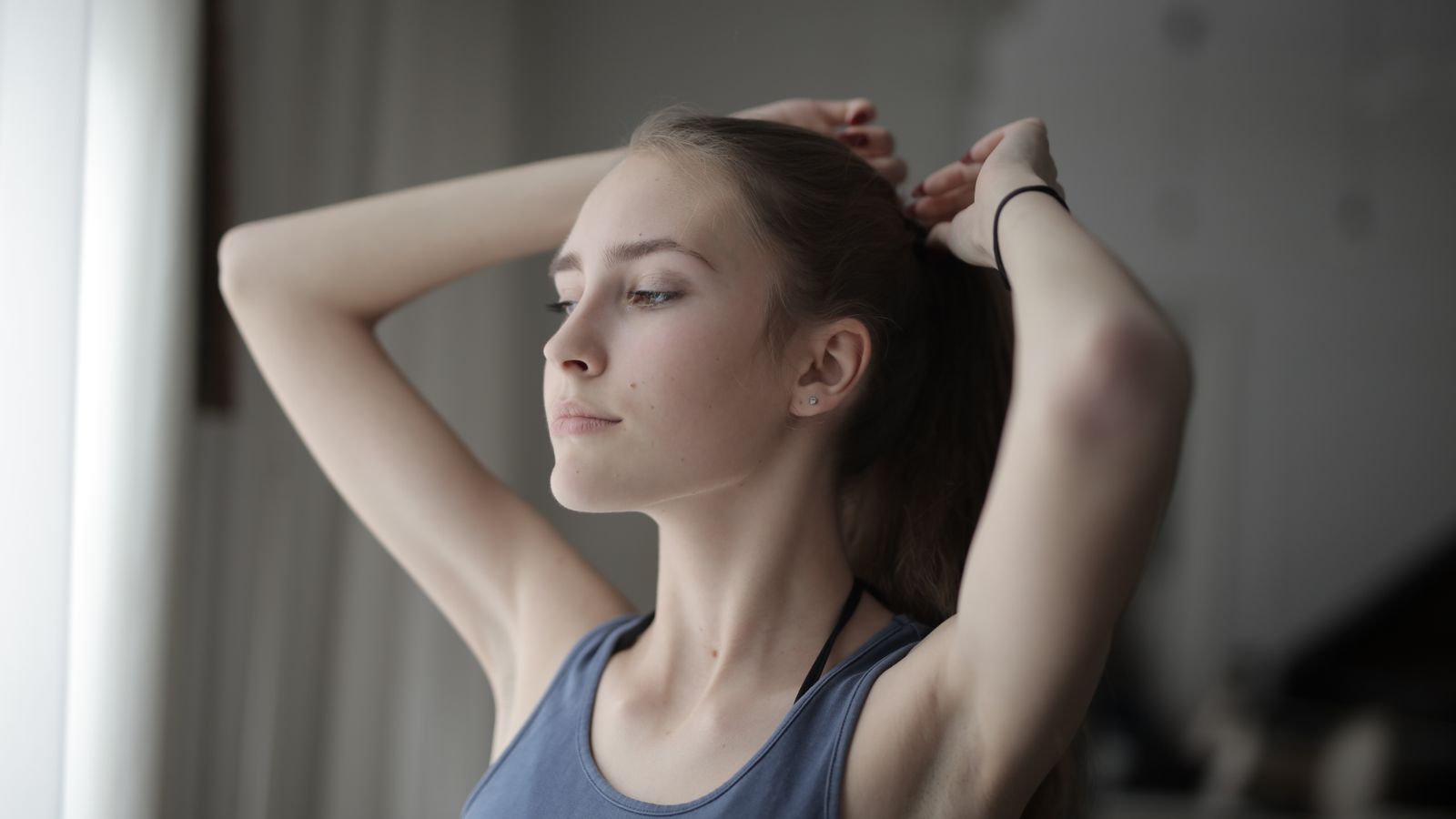Young woman tying her hair up in soft window light, illustrating a low effort hair care routine