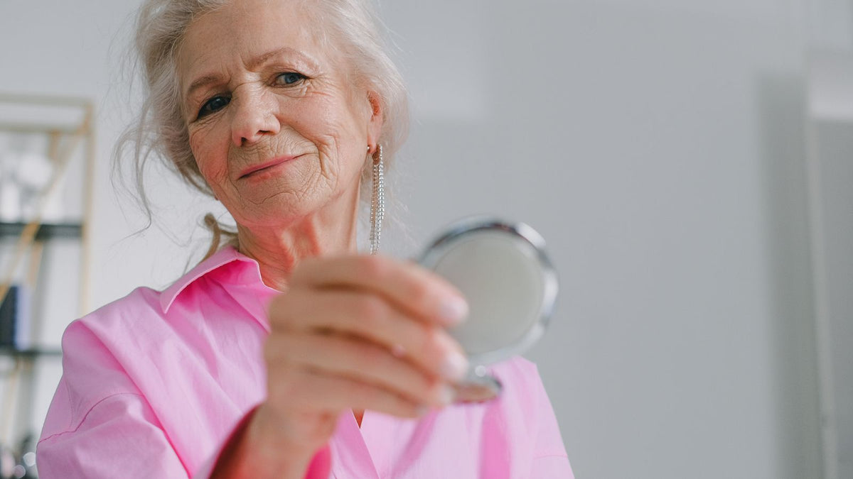 Smiling older woman in a pink shirt holding a compact mirror, checking for menopause dry scalp