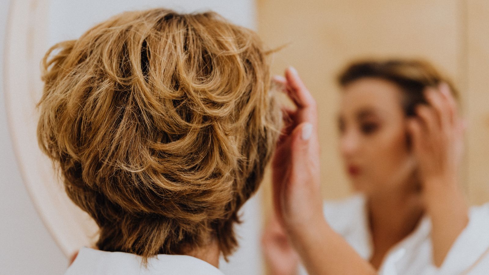 Person styling short hair at a mirror, checking density as transgender hair thinning begins