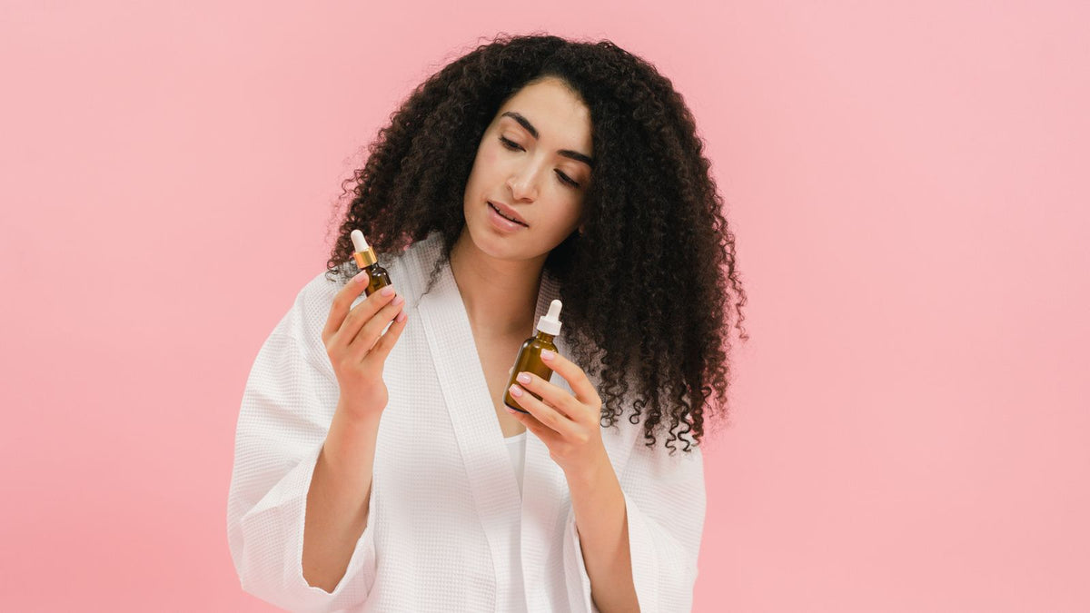 Woman in a white robe comparing two hair oil bottles, the kind of choice readers ask trichologists Gillingham about