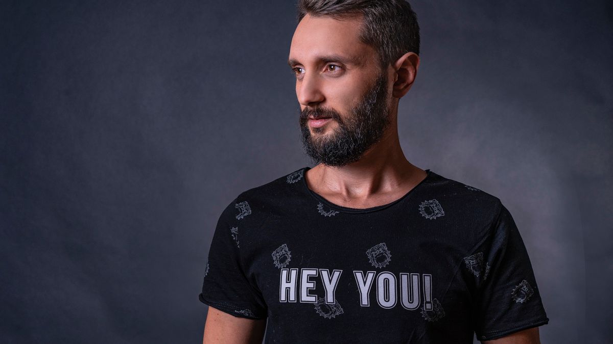 Young man with dark hair and a beard that is turning grey early, photographed against a dark studio background