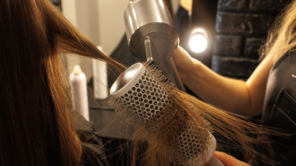 Stylist blow-drying long hair with a round brush, the volume question trichologists Stafford hear often